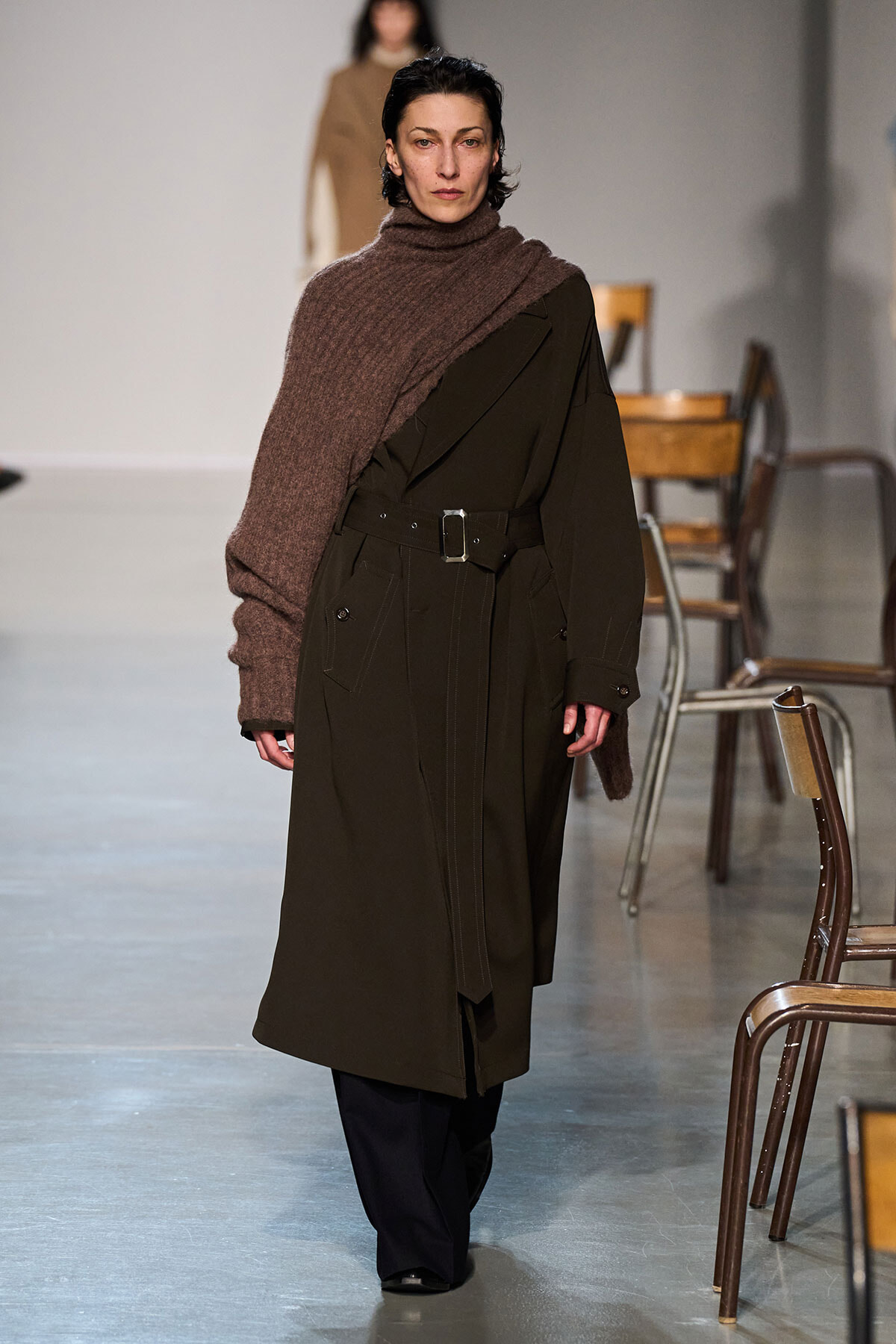 Model walking runway in a dark olive trench coat with a wide brown scarf draped over one shoulder.