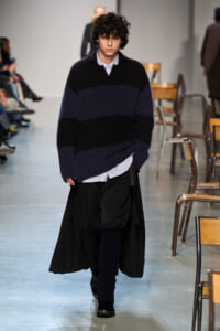 Male model on a runway wearing a chunky navy and black striped sweater over a striped shirt, with a long black pleated skirt and dark socks.