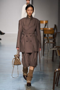 Model walking runway in taupe layered trench coat with belted waist, matching shorts, and brown shoes; carries beige drawstring bag; ribbed tan socks with shoes; neutral backdrop with chairs.