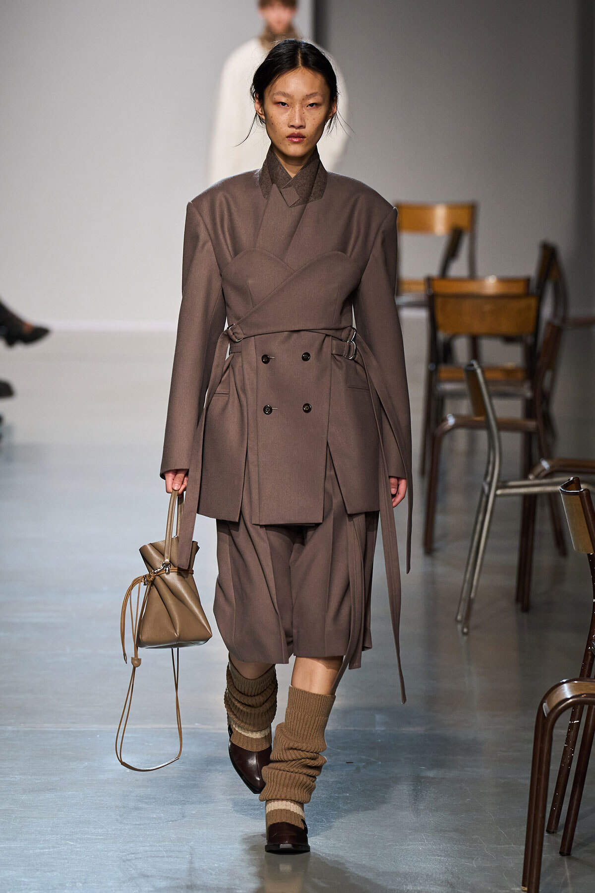 Model walking runway in taupe layered trench coat with belted waist, matching shorts, and brown shoes; carries beige drawstring bag; ribbed tan socks with shoes; neutral backdrop with chairs.