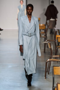 Female model walking a runway in a light blue pinstripe belted trench coat dress with wide lapels and black boots.