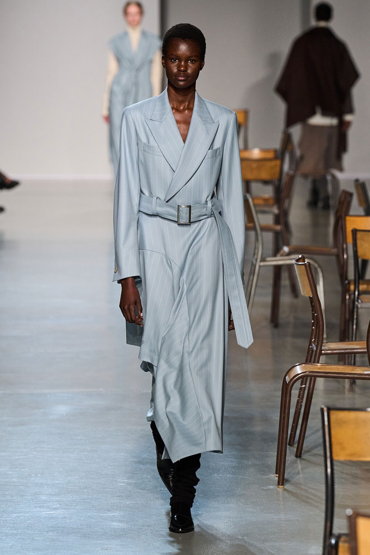 Female model walking a runway in a light blue pinstripe belted trench coat dress with wide lapels and black boots.
