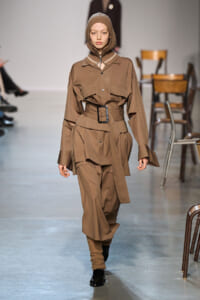 Model on a runway wearing a tan safari-inspired outfit with a hood, wide belt, and layered pockets.