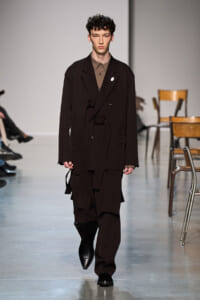 Model walks the runway in a dark brown, oversized layered suit with a belt and long, draped panels, backdrop with chairs visible nearby.