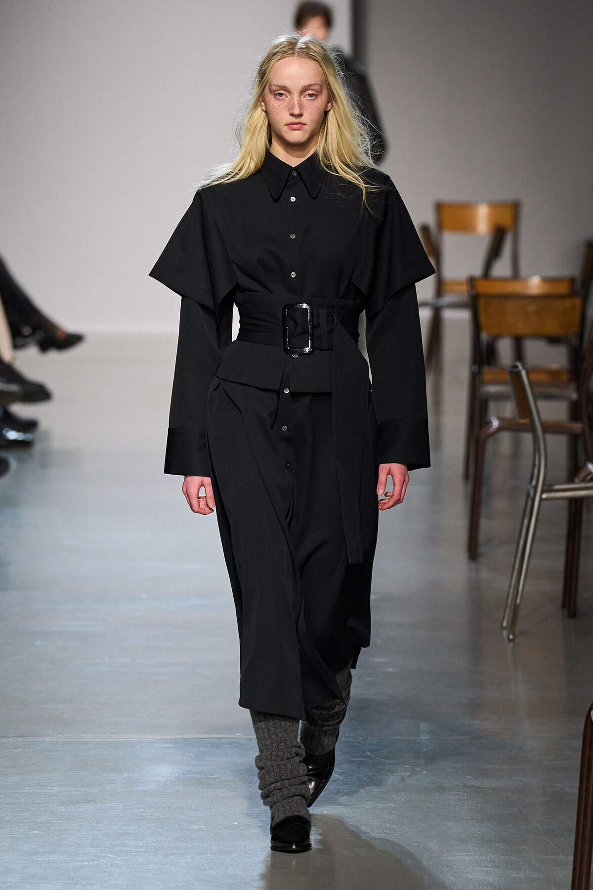 Model walking a runway in a black belted shirt-dress with oversized cape-like sleeves and a wide belt.