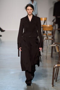 Female model walks the runway in a dark brown, layered trench-style outfit with a waist belt and flowing skirt panels.