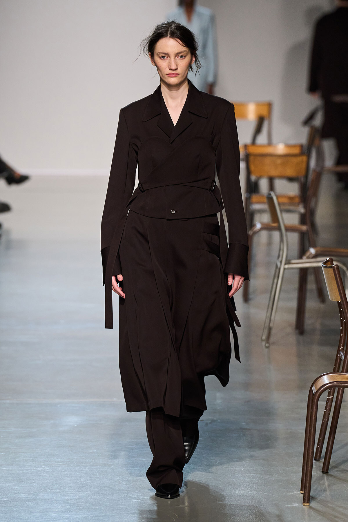 Female model walks the runway in a dark brown, layered trench-style outfit with a waist belt and flowing skirt panels.