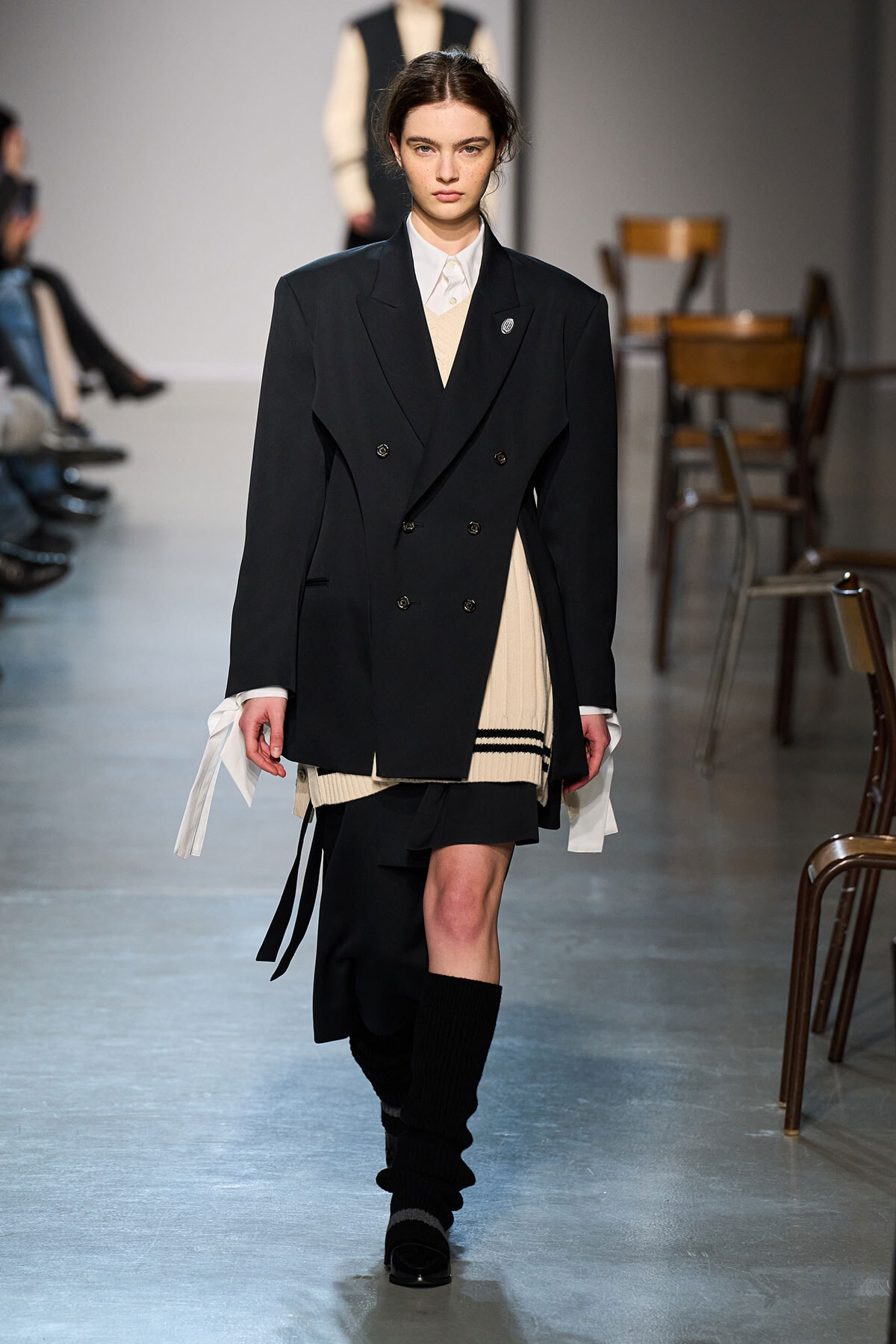 Model walking a fashion runway in a double-breasted black blazer over a cream sweater and white collared shirt, with a beige knit layer and black skirt visible at the bottom.