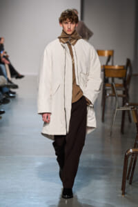 Model on a runway wearing an oversized cream coat over a brown knit turtleneck and scarf, paired with dark brown trousers and black shoes.