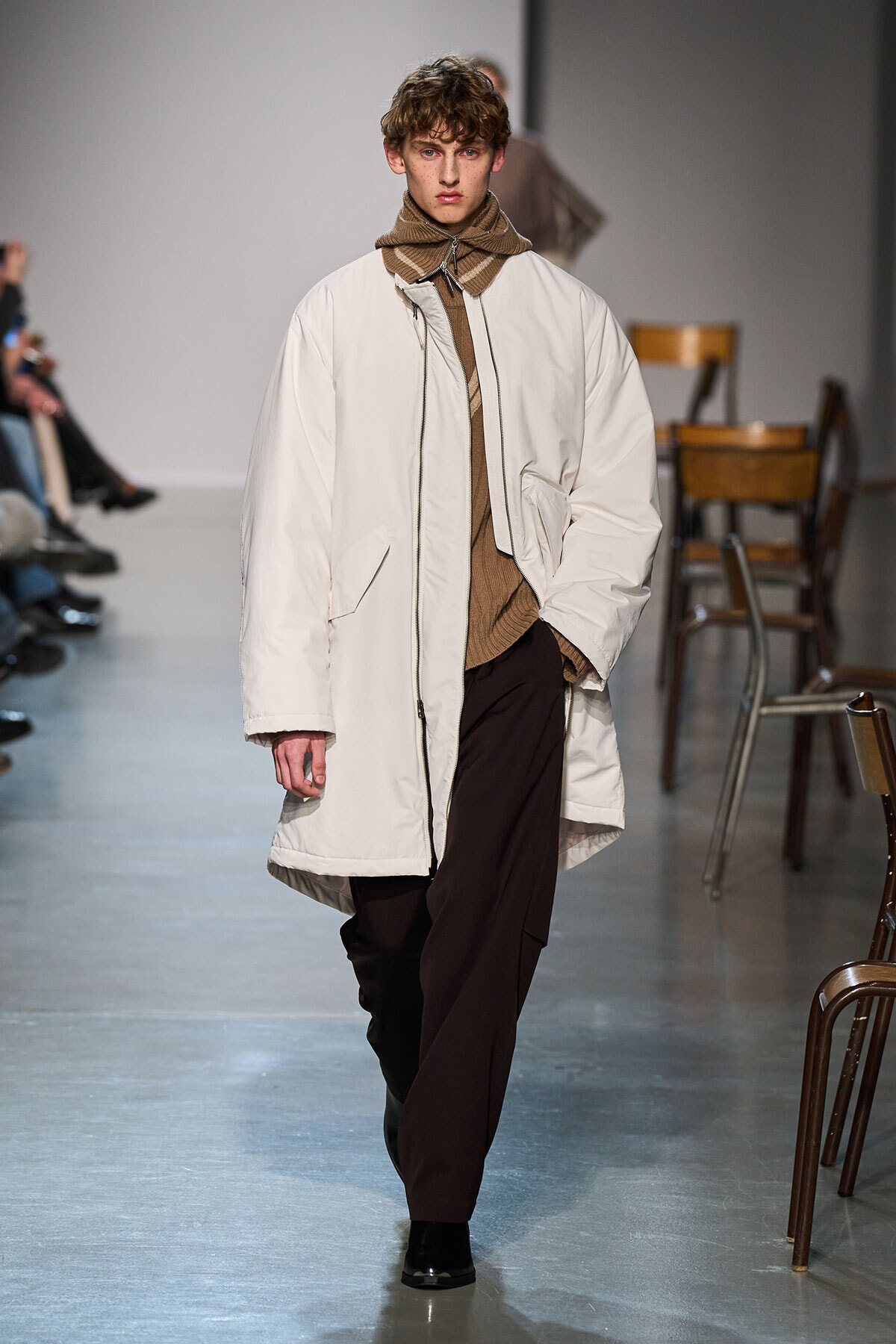 Model on a runway wearing an oversized cream coat over a brown knit turtleneck and scarf, paired with dark brown trousers and black shoes.