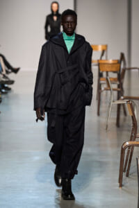 Runway model strides forward in an oversized dark pinstripe blazer over a teal turtleneck and baggy black pants, chairs along the catwalk behind.
