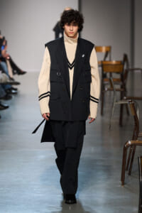 Male model walks the runway in a cream turtleneck under a black utility vest and wide-leg black pants.