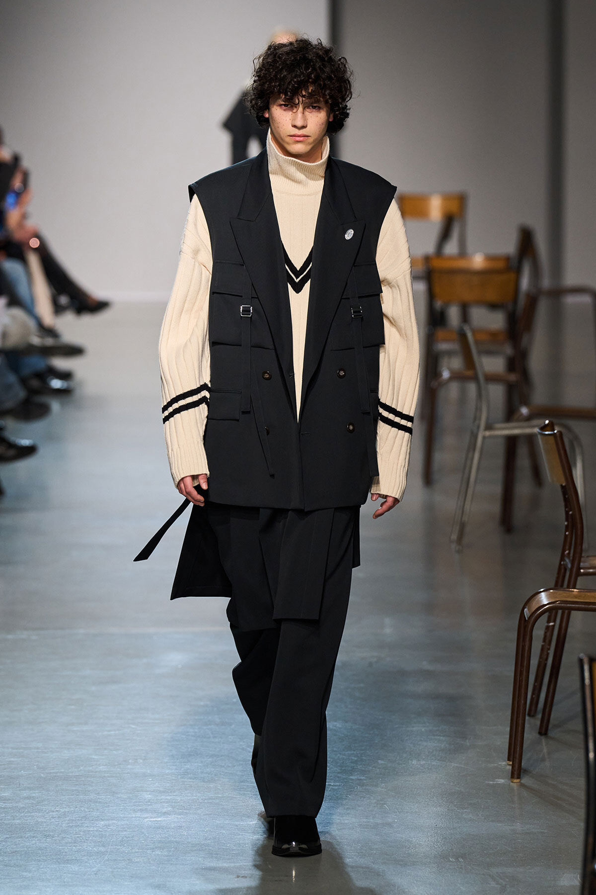 Male model walks the runway in a cream turtleneck under a black utility vest and wide-leg black pants.