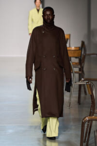 Model walking the runway in a knee-length dark brown double-breasted coat over pale green wide-leg trousers, black boots visible at the feet.