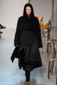 Model walking a runway in an all-black outfit: a fuzzy hooded coat over a quilted, voluminous skirt.