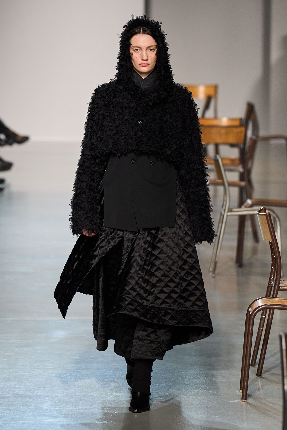 Model walking a runway in an all-black outfit: a fuzzy hooded coat over a quilted, voluminous skirt.