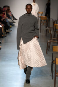 Model walks runway in oversized gray knit turtleneck with beige quilted A-line skirt and gray leg warmers, backstage seating visible on sides.