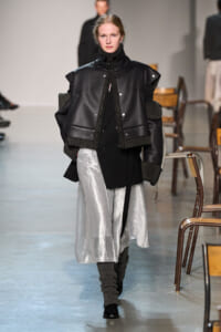 Runway model in a black shearling-trimmed leather jacket over a black outfit with a silver metallic skirt and chunky knit leg warmers.