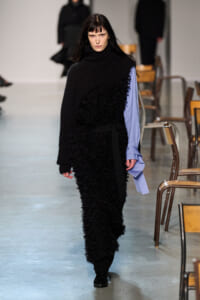 Model walking a runway in a black textured dress with a large knitted scarf and a blue shirt sleeve peeking out