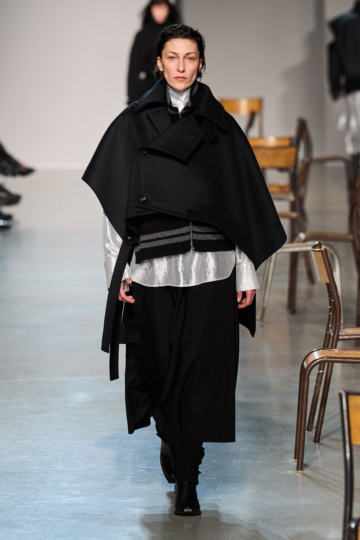 Model walking the runway in an oversized black cape over a metallic silver top and black wide-leg pants.
