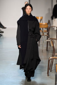 Model walks the runway in a long black dress-like coat with a navy scarf draped over the shoulders