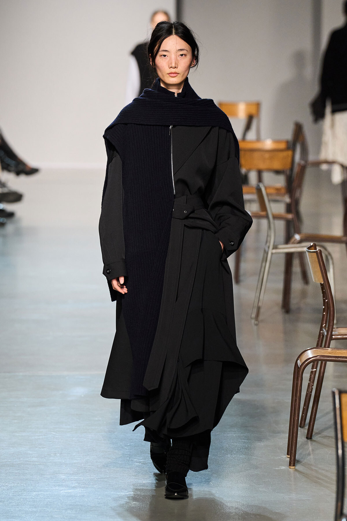 Model walks the runway in a long black dress-like coat with a navy scarf draped over the shoulders