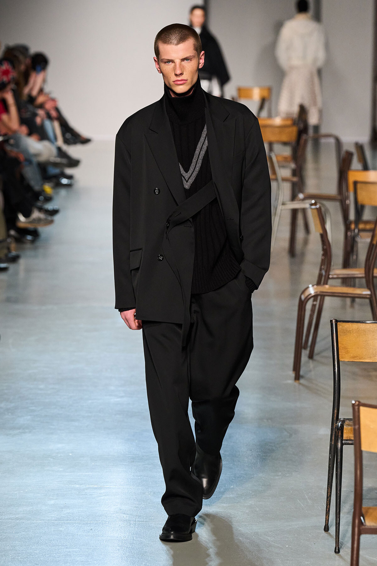 Male model walks the runway in an all-black layered outfit with a long coat and turtleneck over a sweater; audience along the aisle is visible on the left.