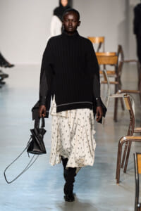 Model walking the runway in a chunky black knit sweater over a white printed skirt, carrying a small black handbag.