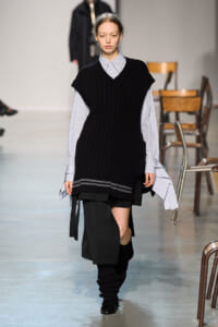 Runway model wearing a black oversized knit vest over a blue striped button-down shirt, with a black asymmetrical skirt and leg warmers.