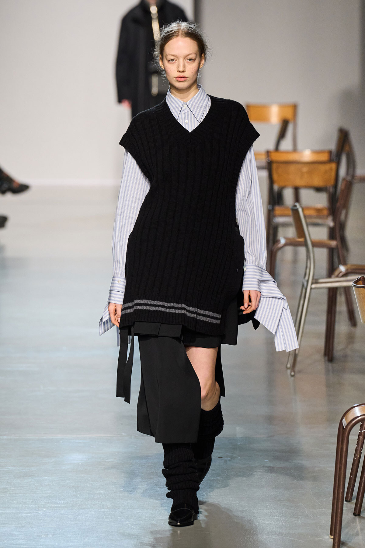 Runway model wearing a black oversized knit vest over a blue striped button-down shirt, with a black asymmetrical skirt and leg warmers.