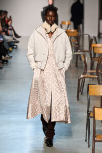 Black female model walks a runway in a pale quilted long coat over a beige dress, with a cream knit collar scarf and hands in pockets to show the outfit’s layers.