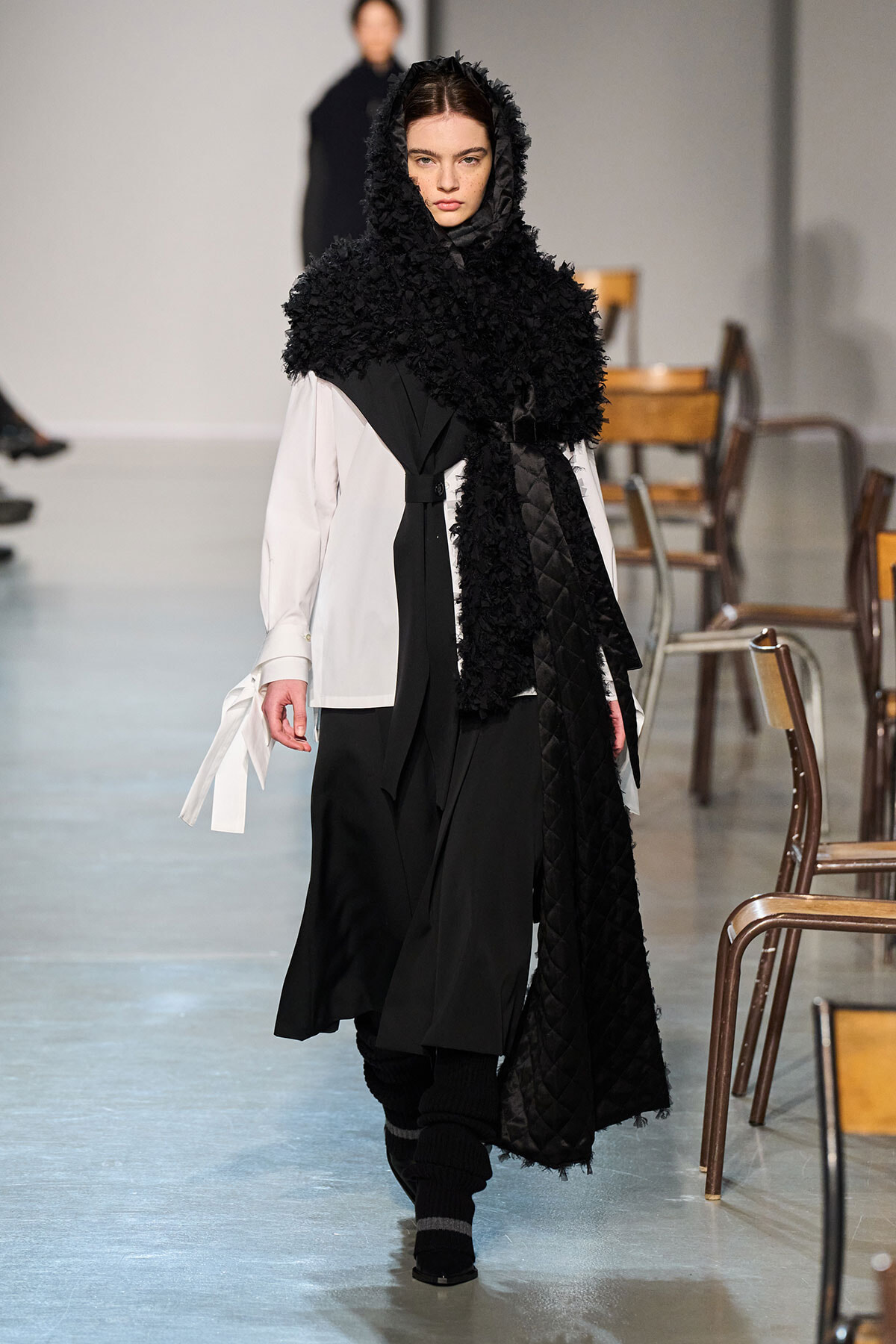 Model walks the runway in a monochrome outfit: white shirt beneath a black layered coat with a large, textured black hood/scarf and quilted drape nearest.