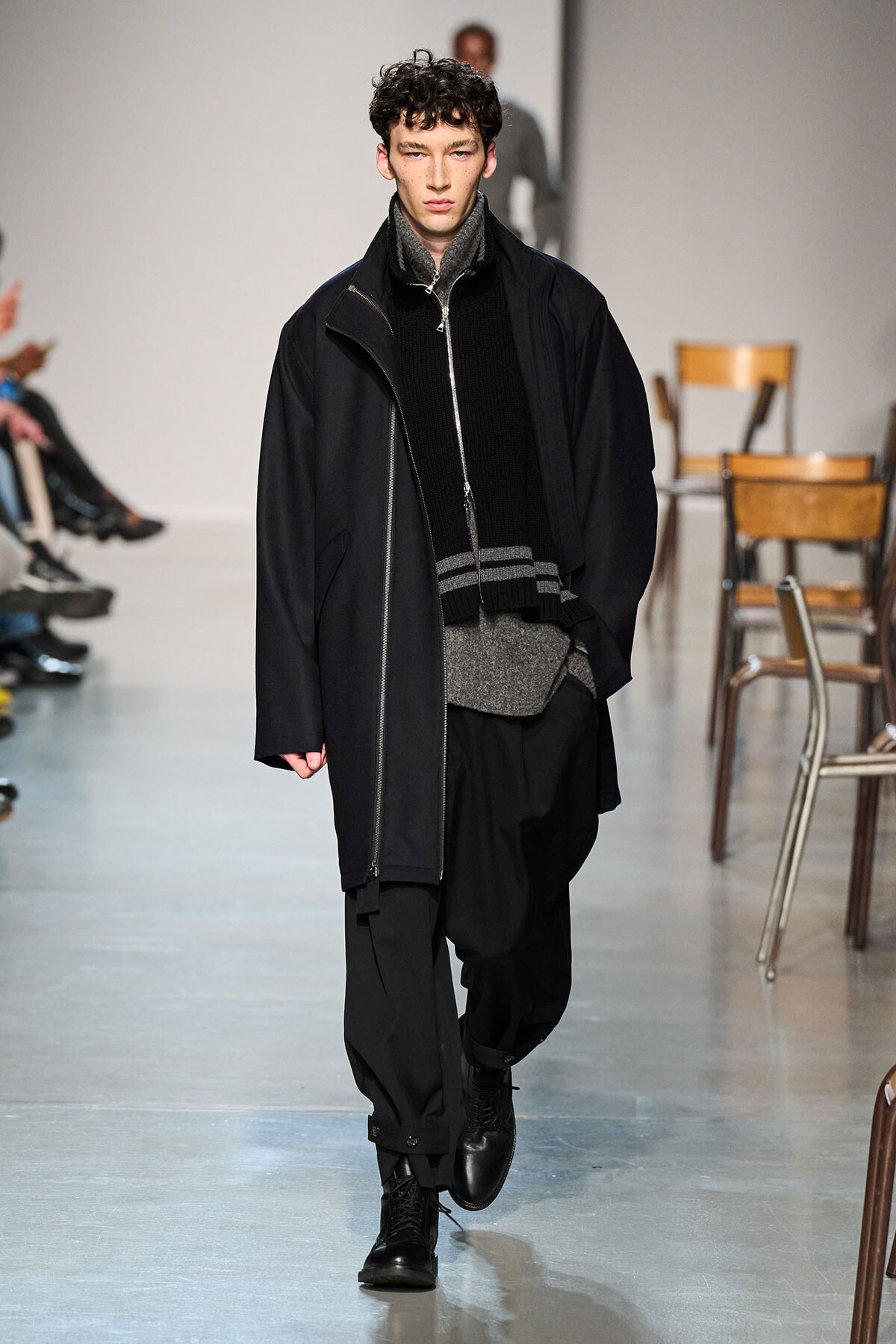 Male runway model in a dark, layered ensemble walking the catwalk; oversized black coat over a zipped jacket, gray knit layers, and black cargo-style pants with black boots
