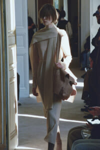 Model walking through a doorway in a beige layered outfit with a draped scarf, carrying a brown coat; audience seated along the hallway.