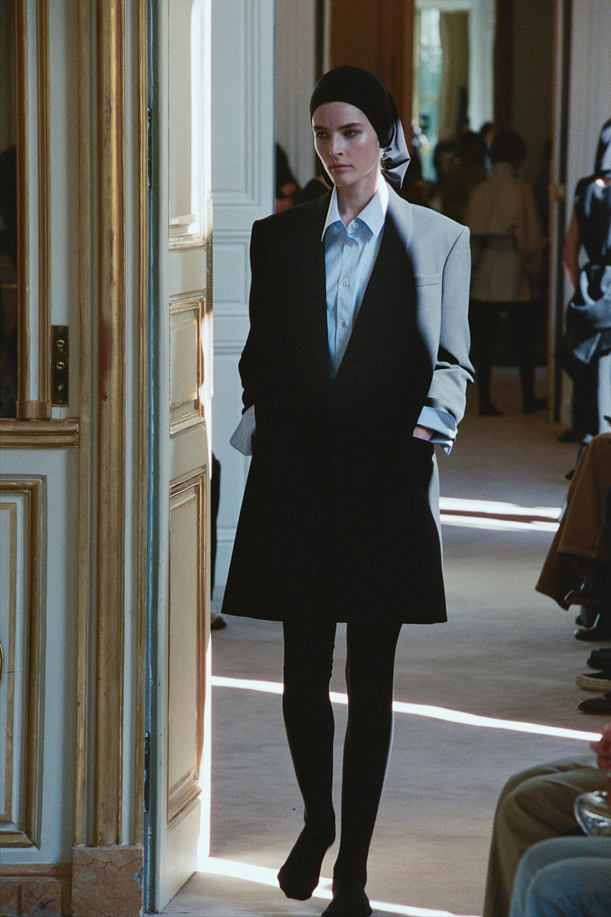 Model wearing a blue collared shirt under a black blazer, black skirt and tights, with a black headscarf, walking through a doorway.