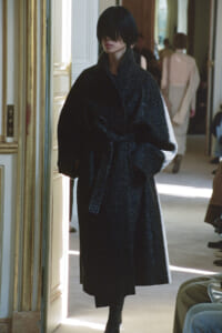 Person in a dark glittery coat walking through a doorway in a fashion show setting, hands in coat pockets, focused expression.