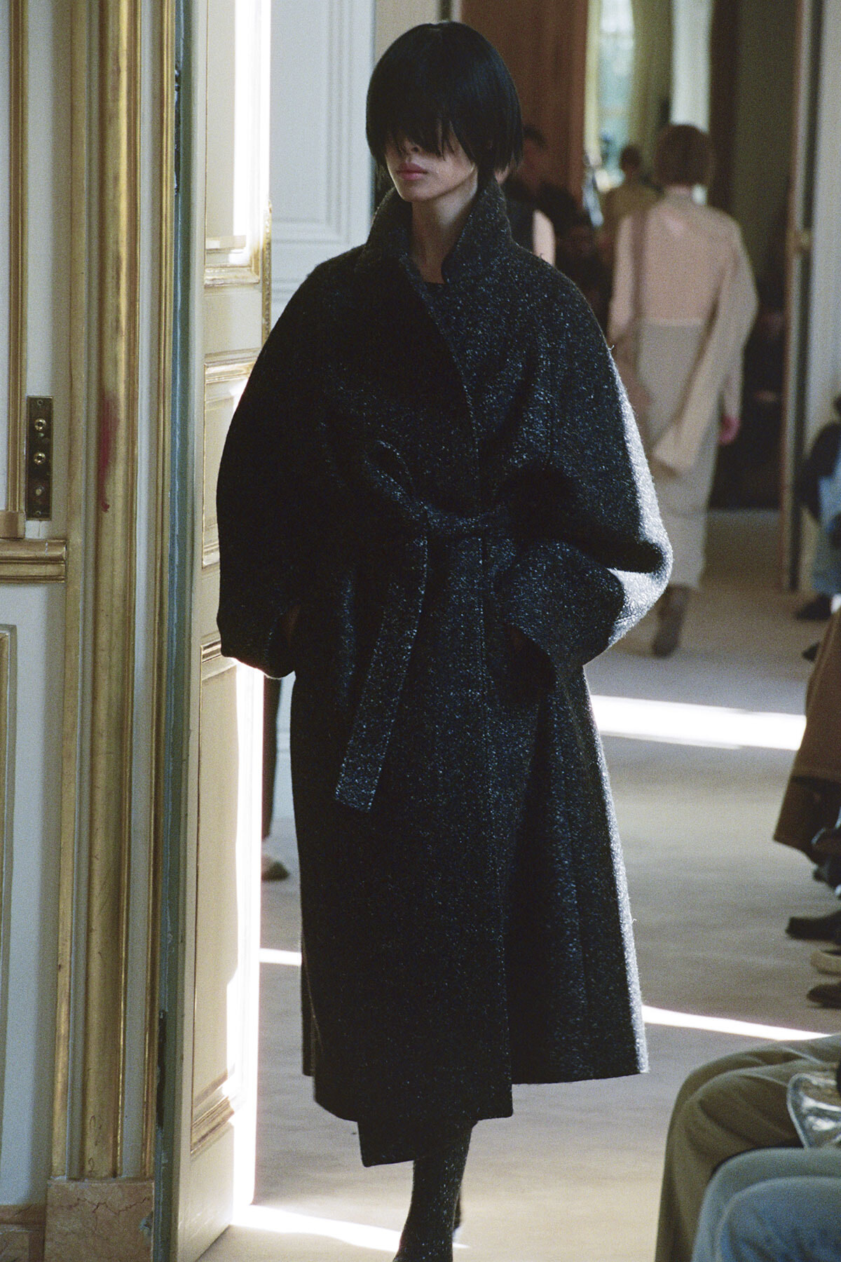 Person in a dark glittery coat walking through a doorway in a fashion show setting, hands in coat pockets, focused expression.