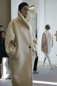 Model wearing an oversized cream wool coat with a high collar on a fashion runway backstage.