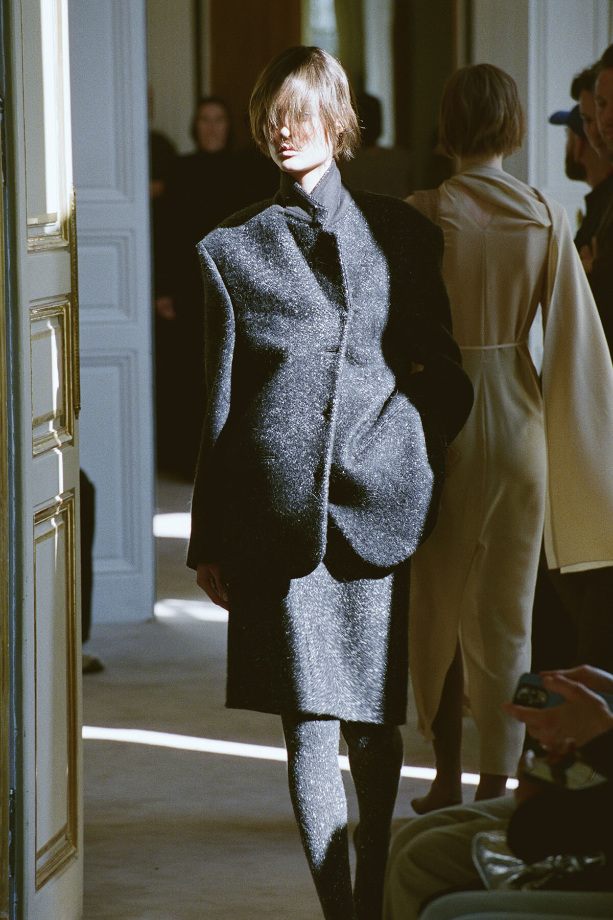 Model in a shimmering charcoal coat and matching skirt walks down a sunlit hallway at a fashion show edge of a doorway visible on the left.