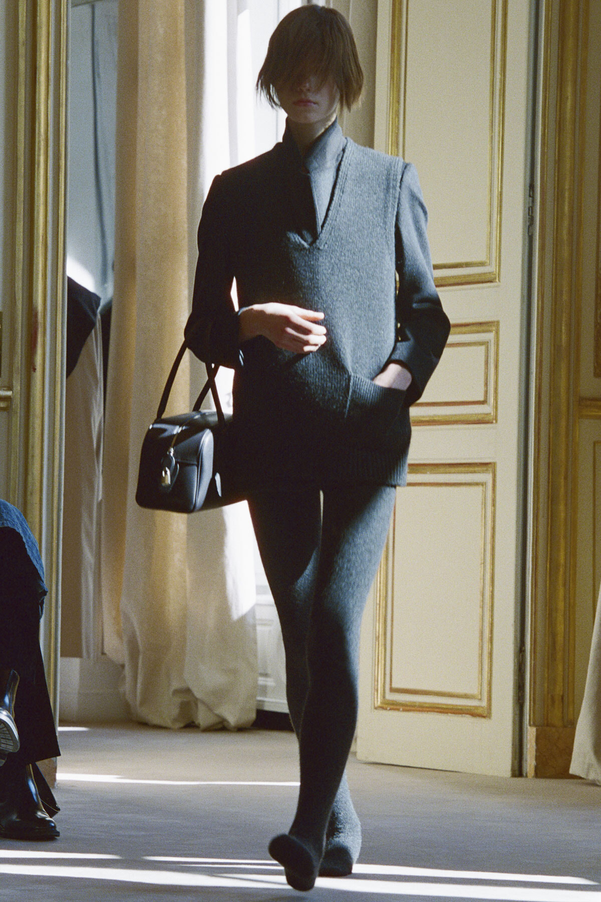 Model in a dark blue sweater dress with matching leggings, carrying a small black handbag in a gilded room.
