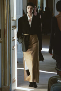 Model walking down a doorway runway in a black blazer, white shirt, and a tan wrap skirt with a slit, wearing a black cap and gloves.