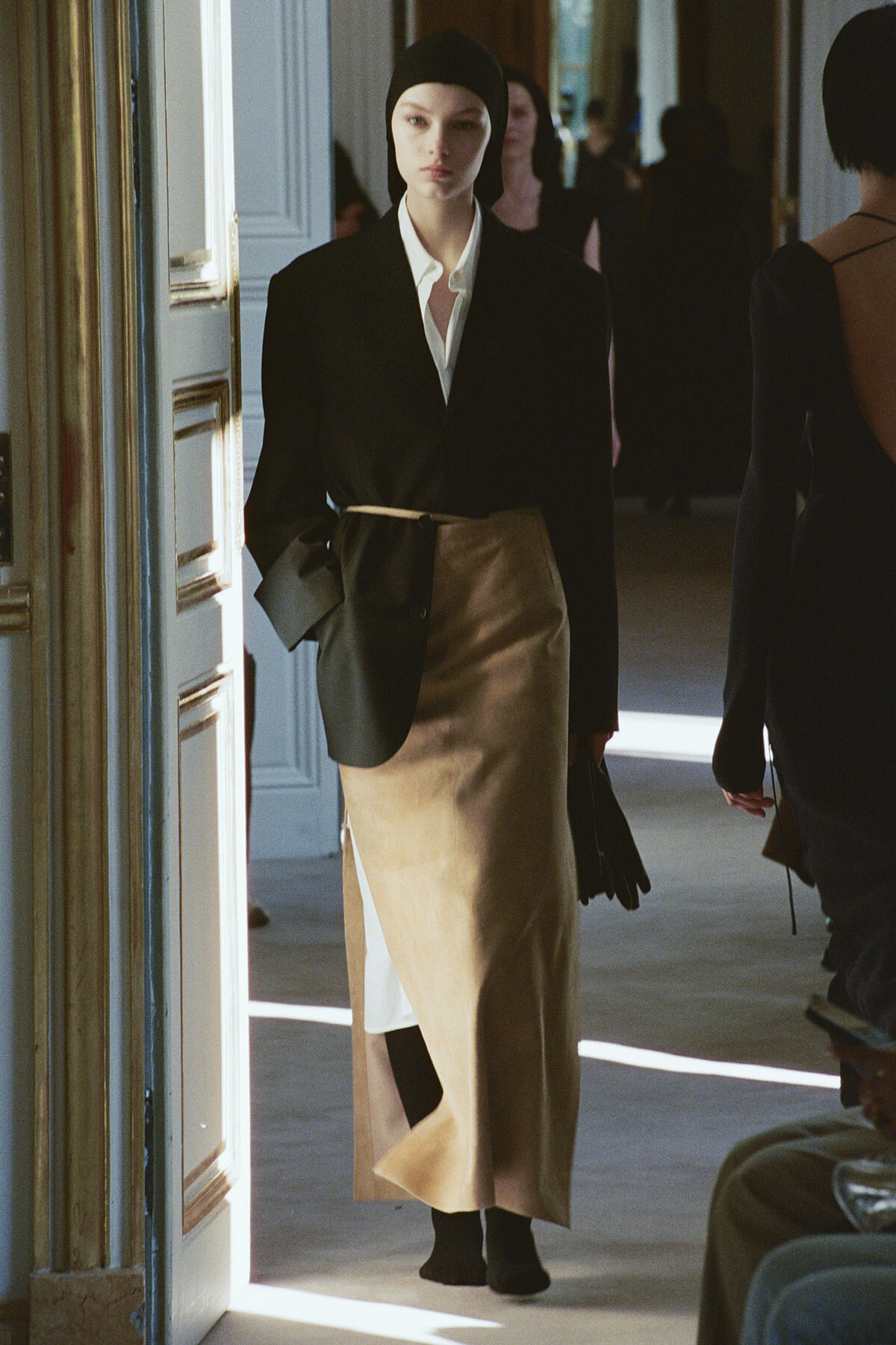 Model walking down a doorway runway in a black blazer, white shirt, and a tan wrap skirt with a slit, wearing a black cap and gloves.