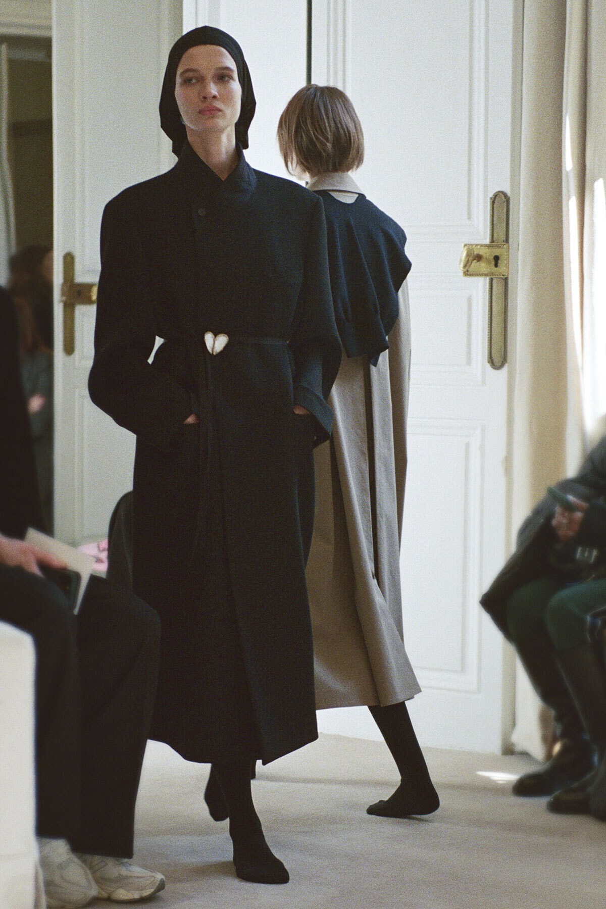 Model walking a runway in a long black coat with a white heart clasp at the waist; another model in a beige cape stands behind in a bright room.