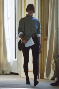 Person walking through a bright interior wearing a knotted gray oversized top, white shirt glow, black leggings, and a small shoulder bag.