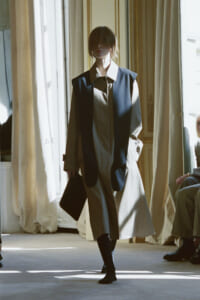 Model walking a fashion show in a long two-tone trench coat with a large white collar, holding a dark clutch as light pours in from windows.