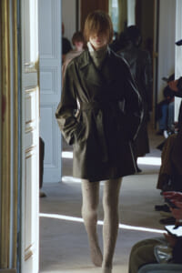 Model walking a fashion runway in an oversized dark coat with a belt and textured beige tights, audience seated along the sides.