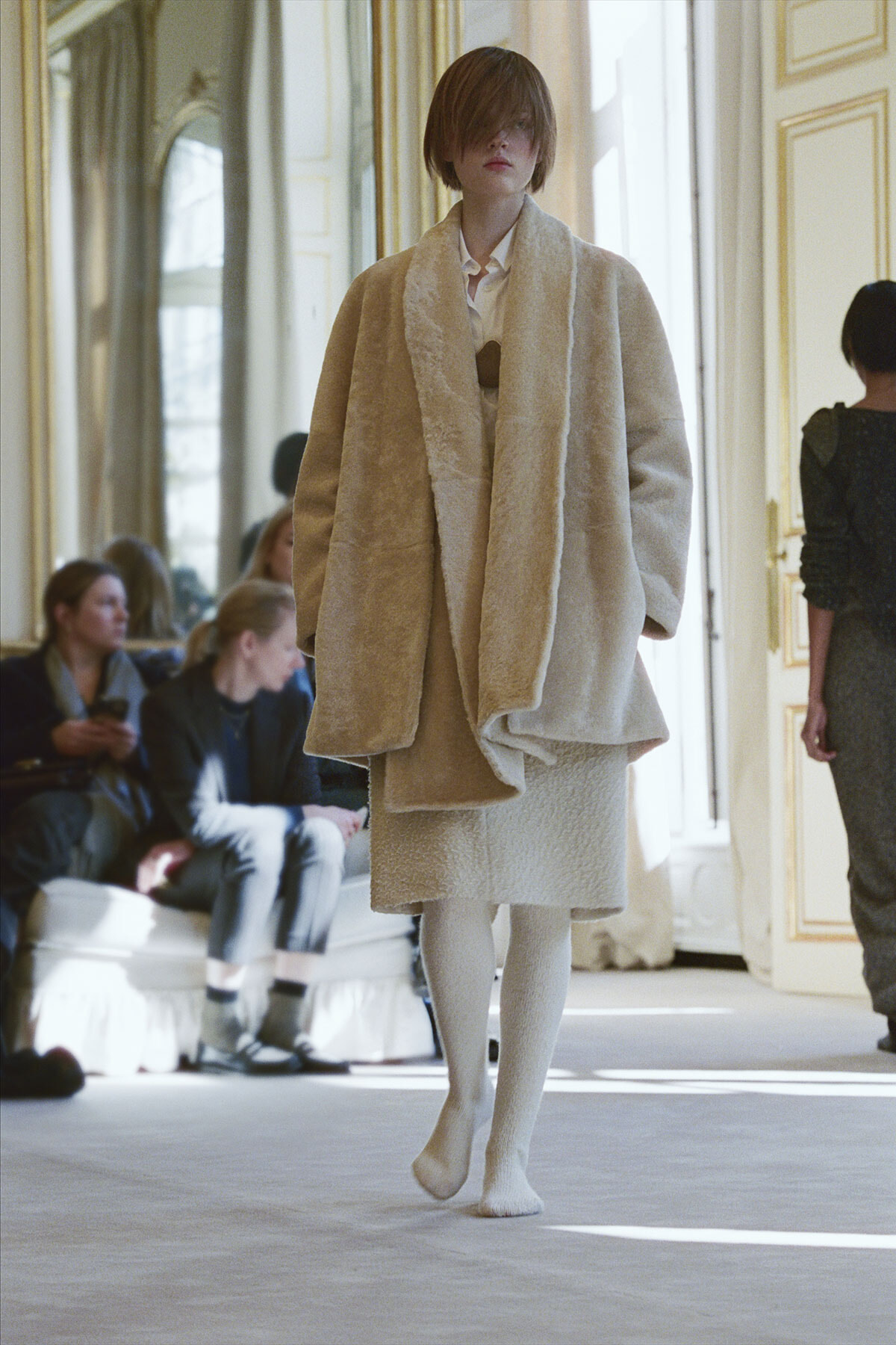 Model walks a runway in a voluminous beige coat with a matching textured skirt and white tights; audience sits in a luxe room in the background.