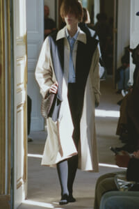 Model walking in a long beige trench with a black color-block front, blue shirt, and a reptile-patterned bag in a bright doorway setting