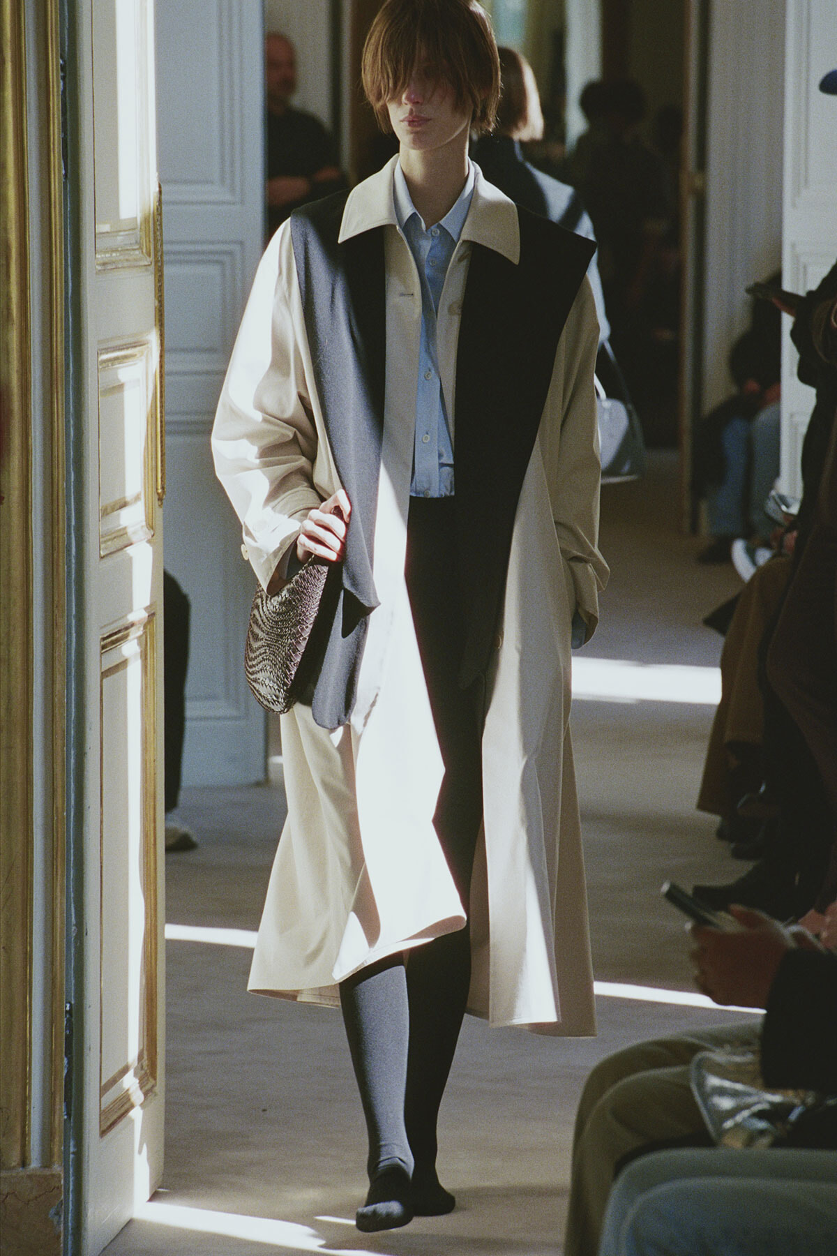Model walking in a long beige trench with a black color-block front, blue shirt, and a reptile-patterned bag in a bright doorway setting