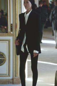 Woman in a black suit and white shirt stands at a gilded doorway, carrying a shoulder bag in a fashion setting.